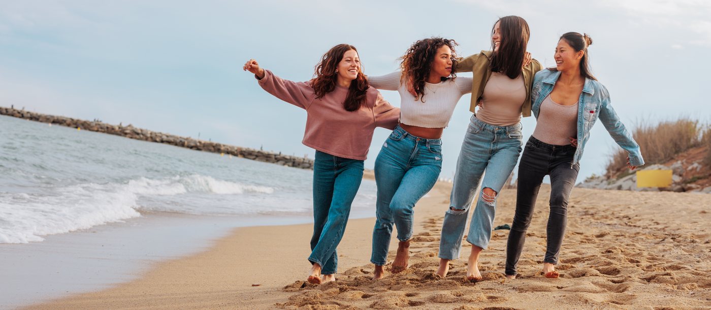 Four friends walking arm in arm along a beach, laughing.