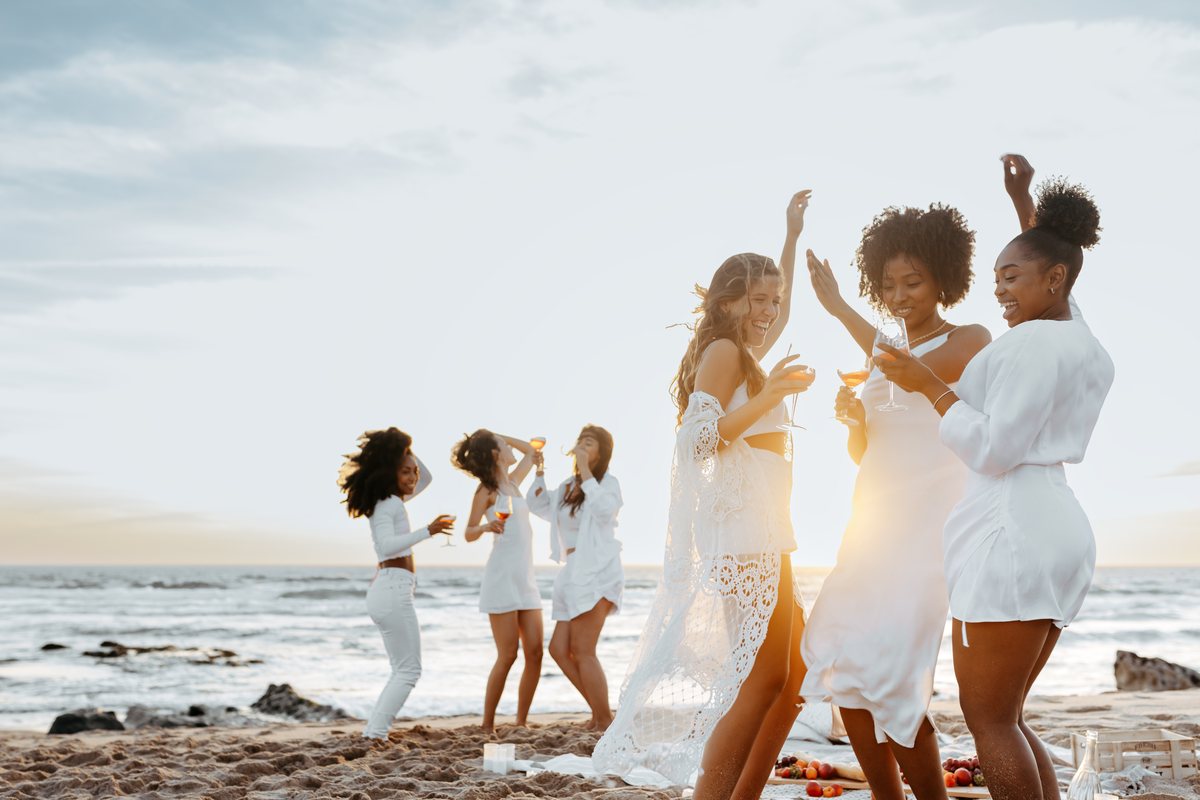 A group of friends in white dancing and toasting on the beach at a bachelorette celebration.